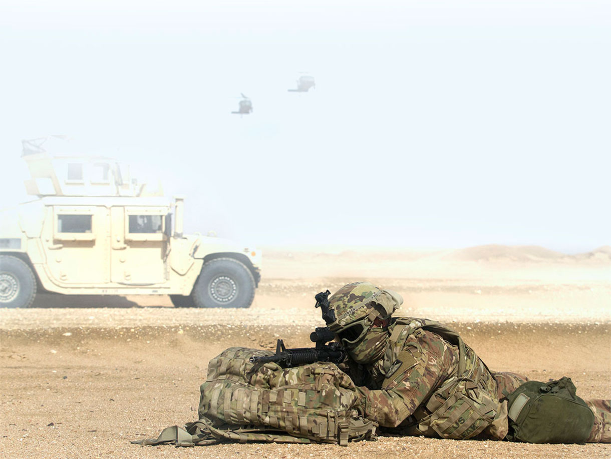 a prone soldier in the desert wearing a mask and goggles, shooting over a backpack, with an armored vehicle in the background